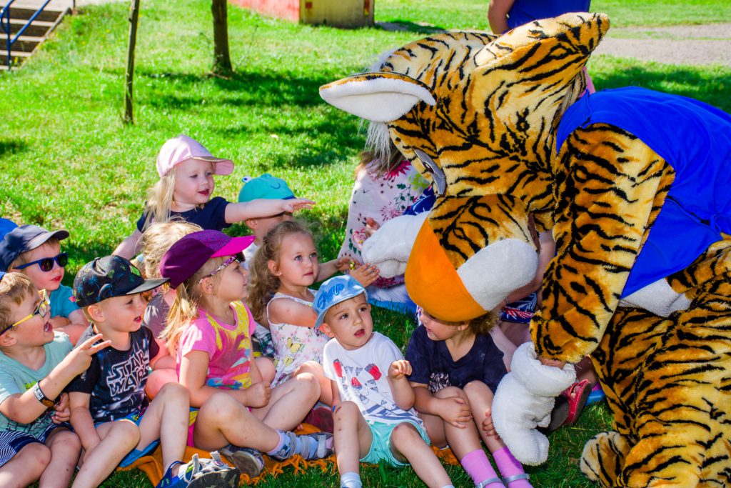 Przedszkolaki siedzące na trawie w towarzystwie wielkiej maskotki - Tygrysa
