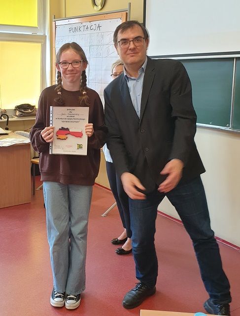 Uśmiechnięta dziewczynka trzyma dyplom za udział w konkursie, stoi razem z nauczycielem