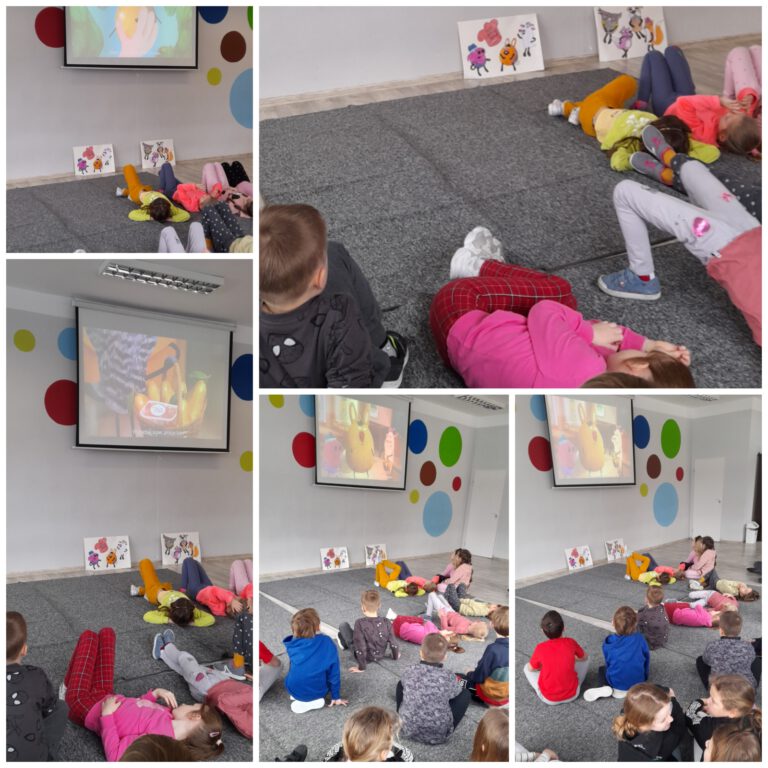 Dzieci siedzą i leżą na dywanie w świetlicy i oglądają prezentację na ekranie projekcyjnym