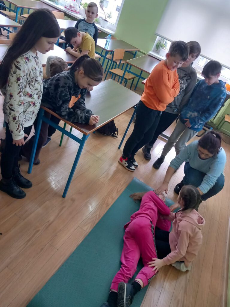 uczniowie ćwiczą pozycję boczną bezpieczną, uczennica leży na podłodze w pozycji