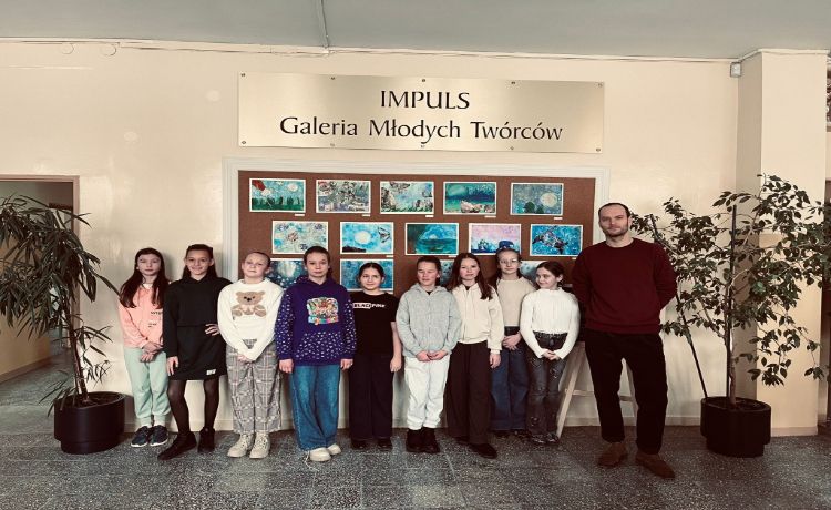 grupa uczniów z nauczycielem stoi na tle galerii prac plastycznych